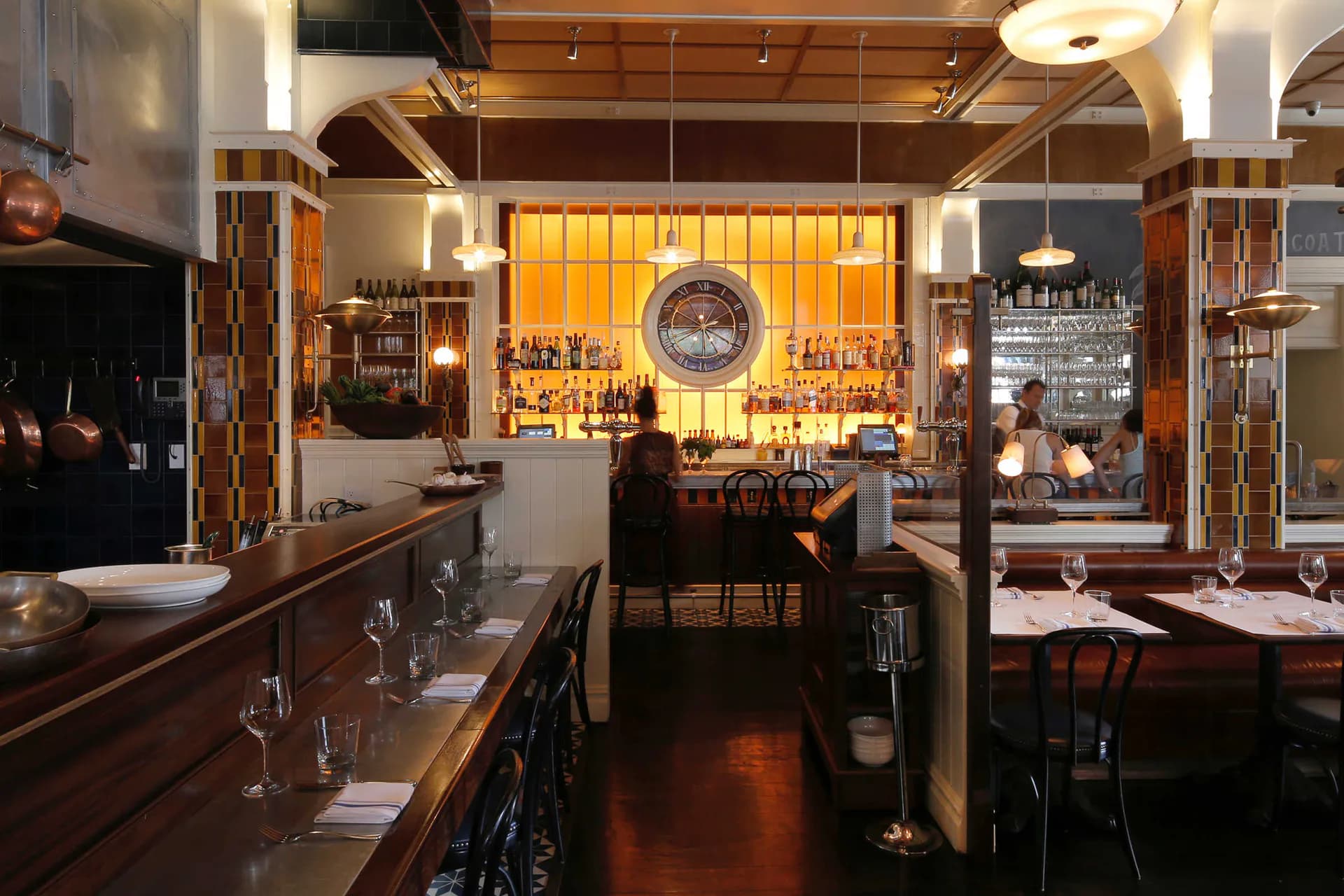 Warm, intimate restaurant interior with wooden ceiling, amber-lit bar display, dark wood furnishings, and glowing window backdrop by Hervé Descottes.