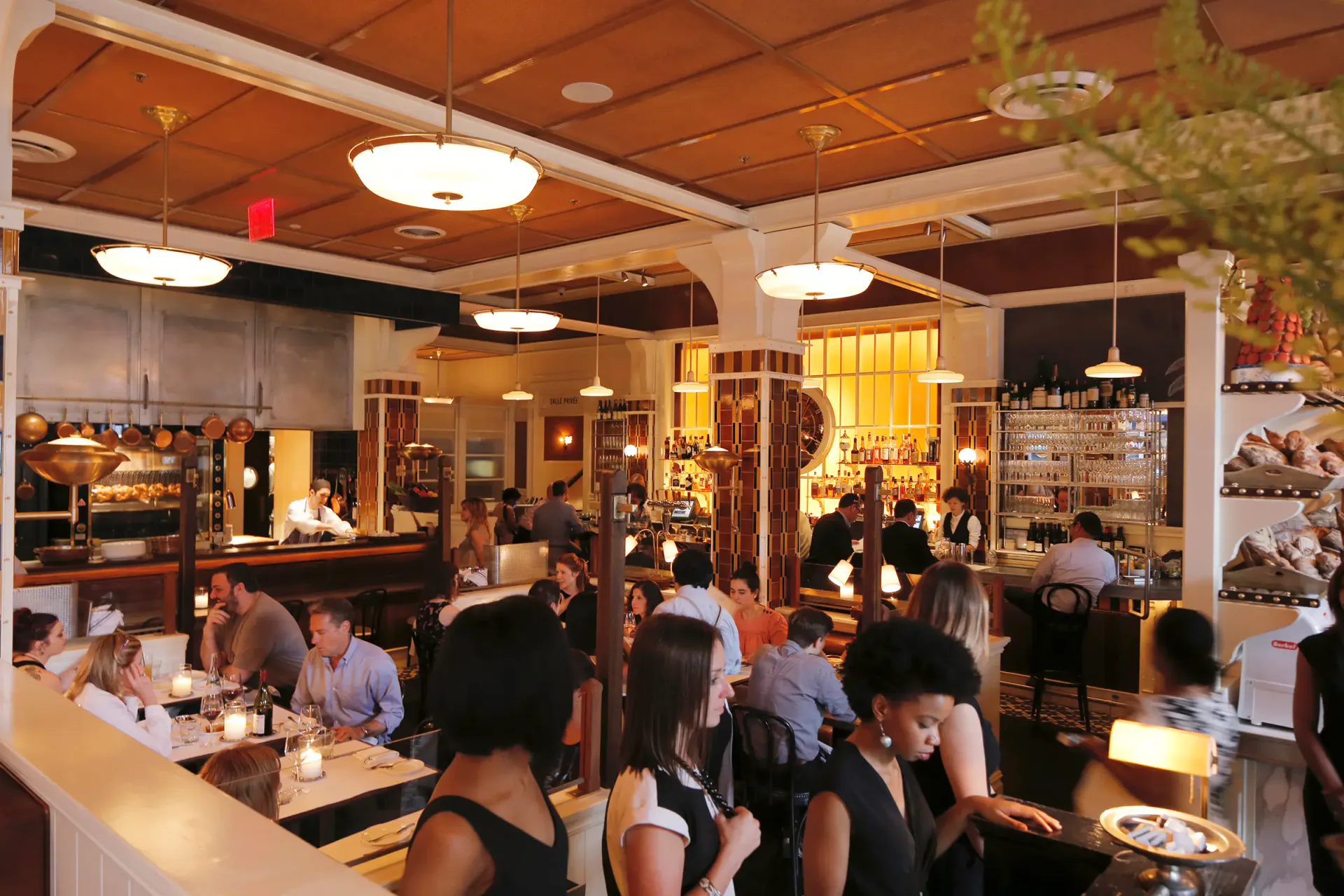 A bustling modern restaurant interior with warm wooden ceiling, pendant lighting, and an open kitchen concept with customers dining and interacting, designed by Hervé Descottes.