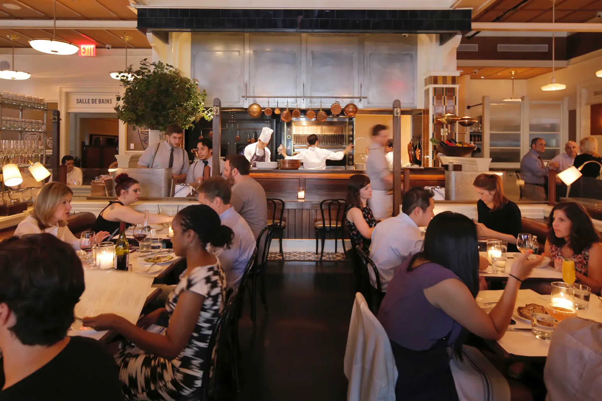 A warm-lit restaurant interior with wooden ceiling details, communal dining tables, and an open kitchen visible in the background, designed by Hervé Descottes.