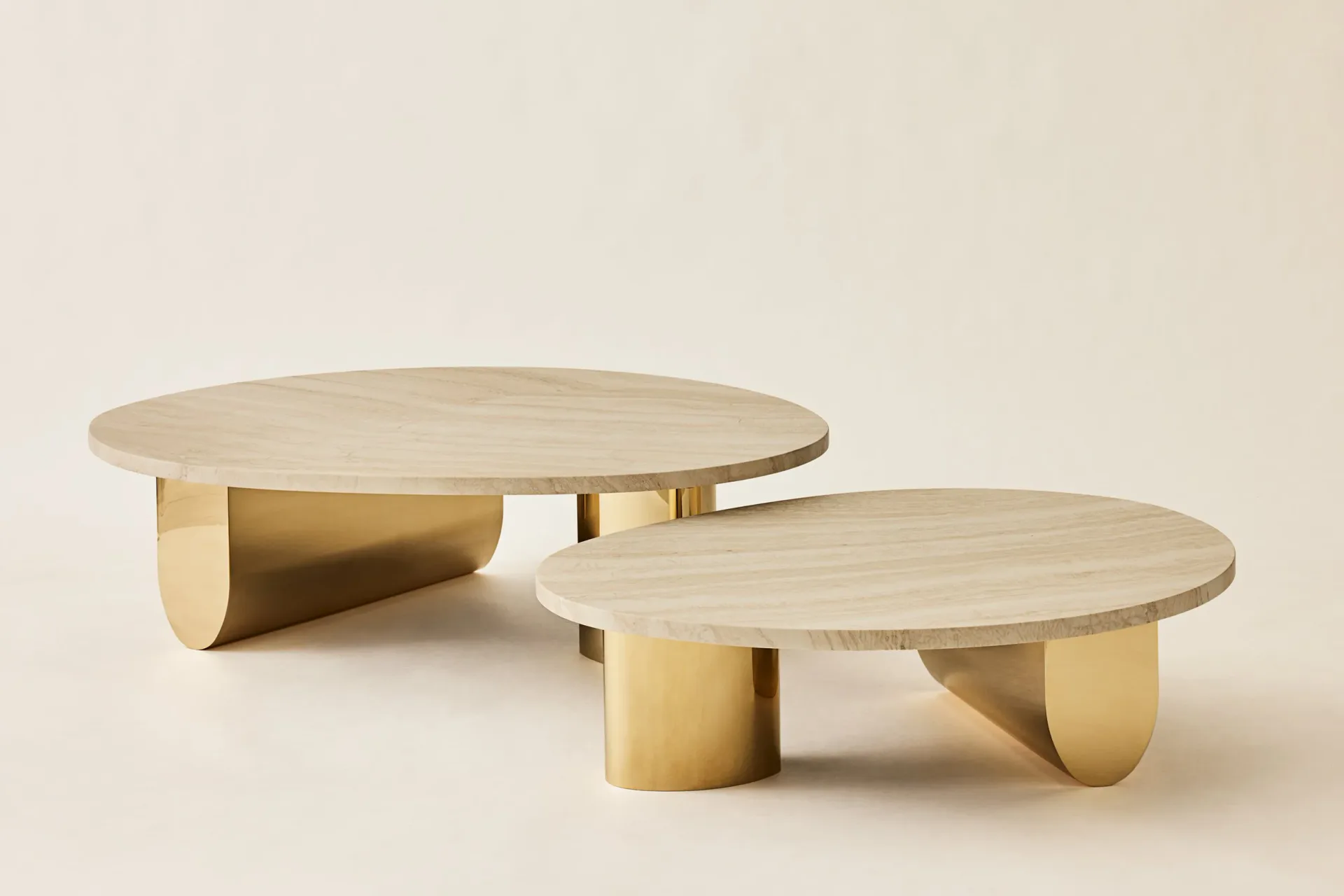 Two sculptural brass and travertine coffee tables with organic curved forms and cylindrical bases on a neutral background by Hillary Petrie.