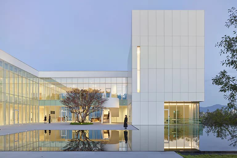 Modern institutional building with white paneled facade, glass curtain walls, reflecting pool, and minimalist landscape design by Hiuki Lam.