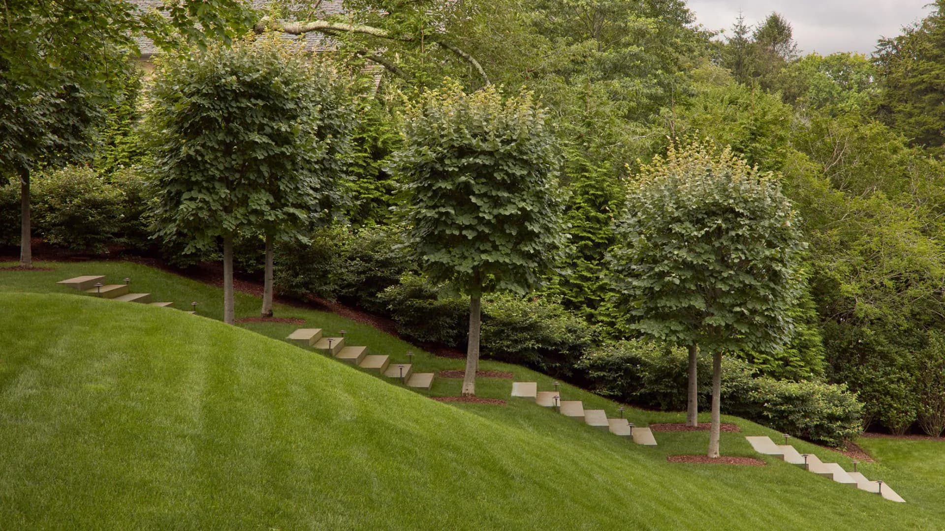 Sloped lawn with geometric stone stepping stones leading through mature trees and layered hedging, creating a formal landscape composition by Hope Brice.