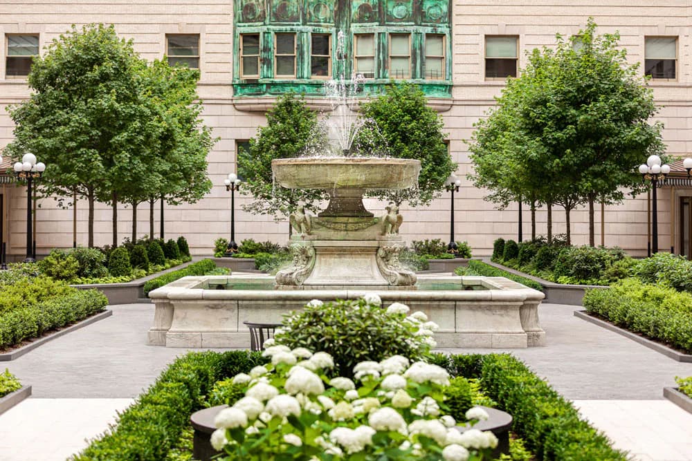 Formal courtyard featuring symmetrical landscaping with white hydrangeas, manicured hedges, topiary trees, stone fountains, and classical architecture by Hope Brice.