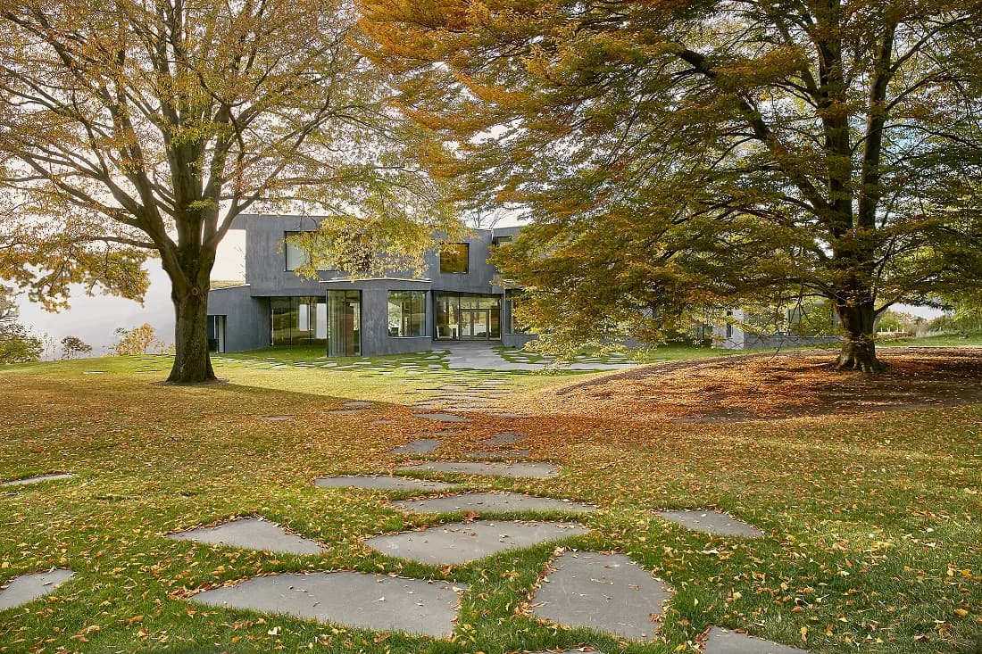 Modern gray residential building nestled among mature trees with stone stepping-stone pathway across leaf-covered lawn by Hope Brice.