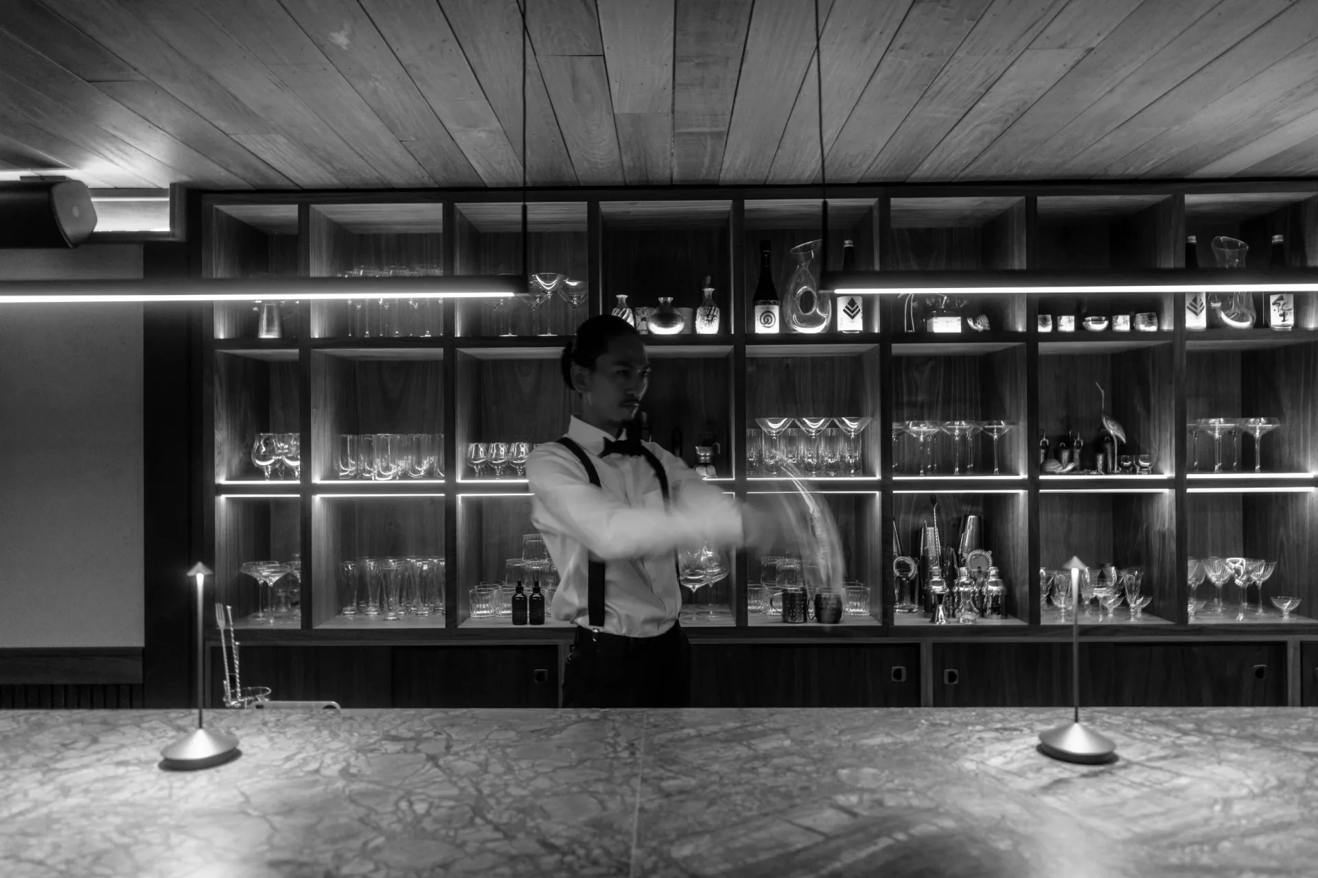 Modern black and white interior with illuminated display cabinets containing glassware and objects, polished concrete floor, and geometric wooden ceiling by Ian Bentley.