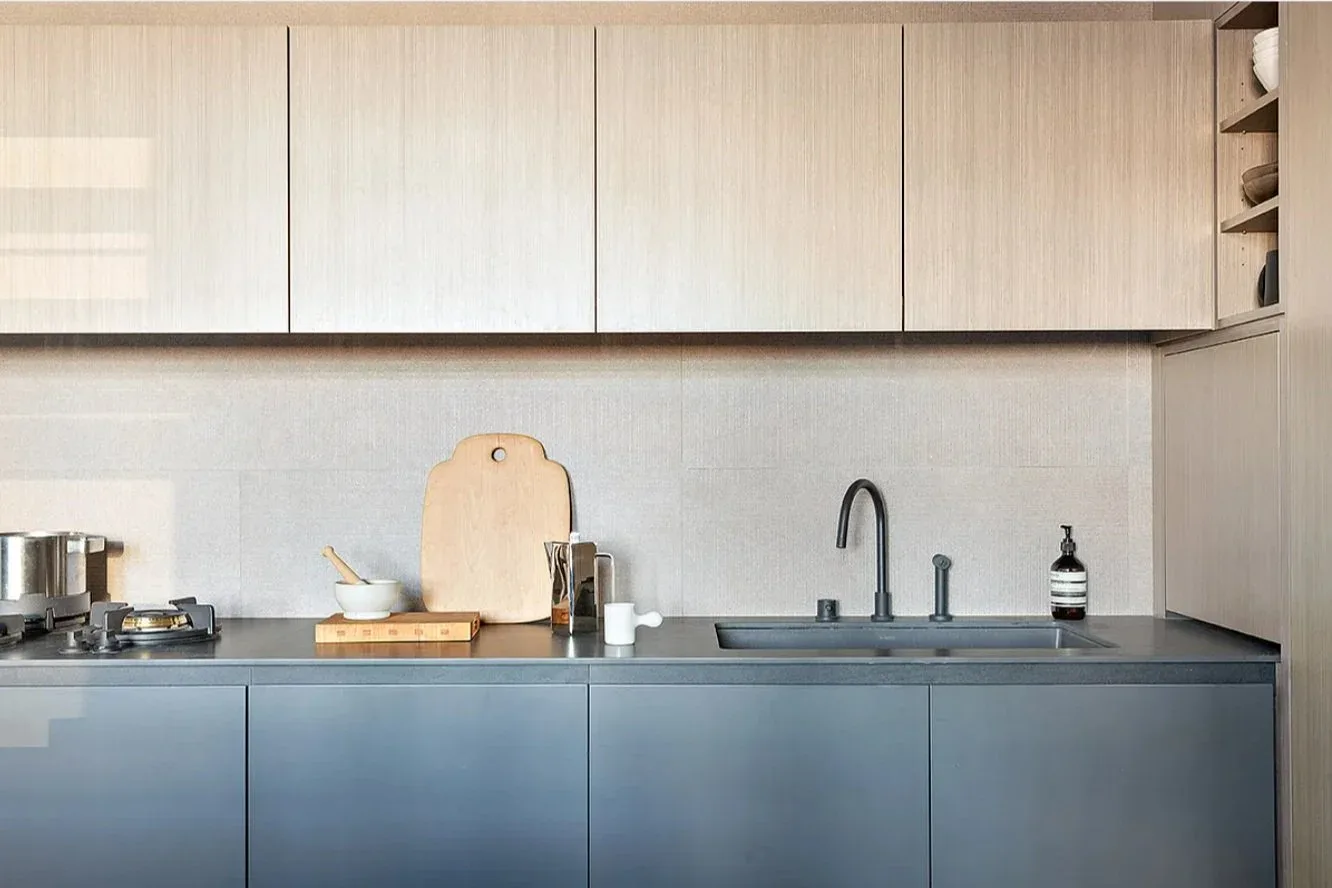 Modern kitchen with light wood upper cabinets and slate blue lower cabinetry, minimalist white backsplash, and integrated sink by Ian Carrington.