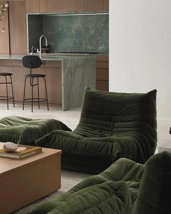 Modern living room featuring olive green textured lounge seating, wooden side table, and kitchen area with green marble backsplash and black bar stools by Ian Carrington.