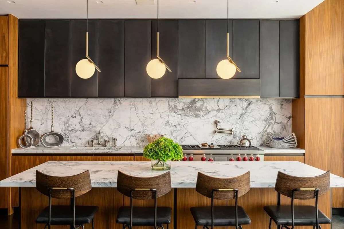 Modern kitchen with marble backsplash, dark upper cabinets, wood lower cabinetry, white waterfall island with brass pendant lights, and upholstered bar seating by Ian Carrington.