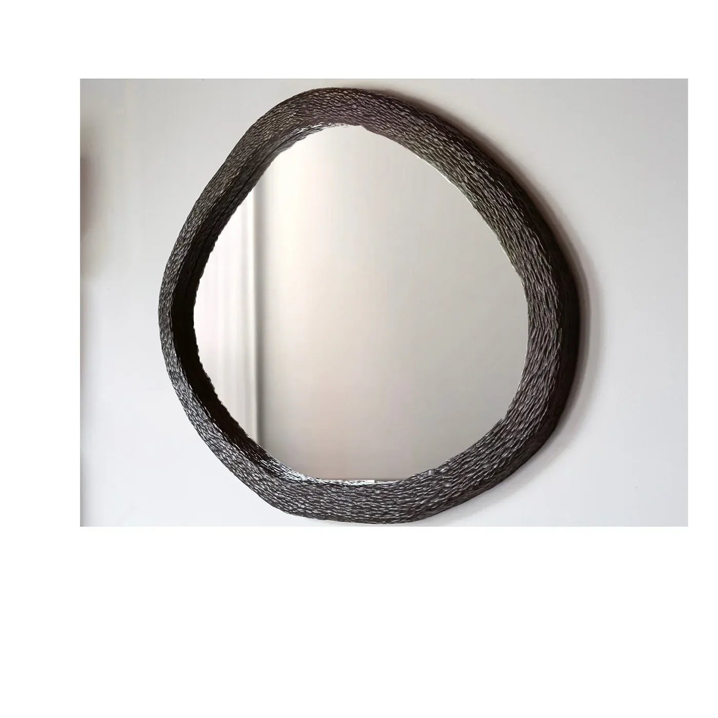 Textured dark circular mirror with organic woven frame mounted on white wall, reflecting light, design by Ian Love.