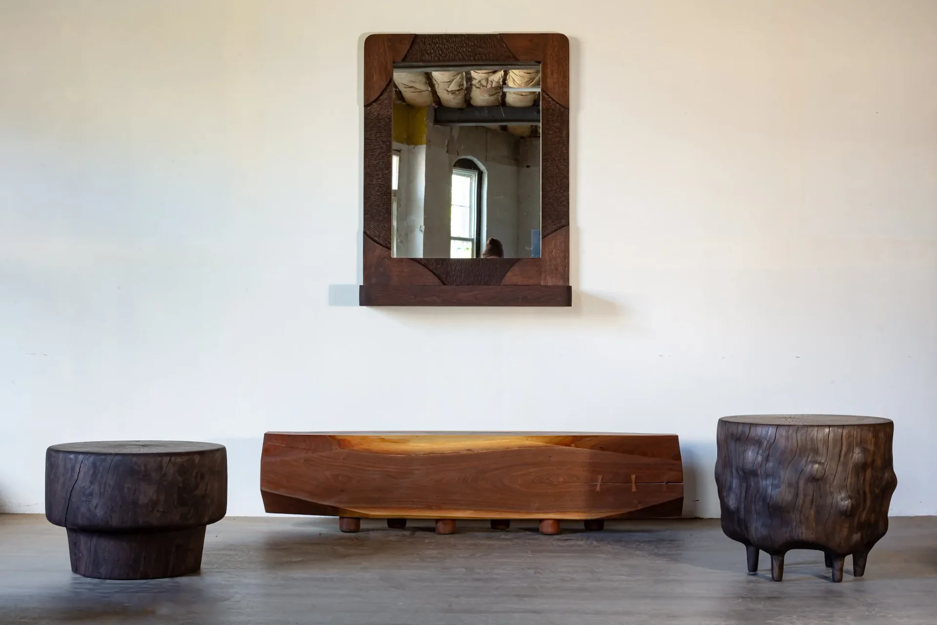Minimalist interior with dark wooden furniture pieces including cylindrical stools and low bench, complemented by wall-mounted mirror, neutral palette by Ian Love.