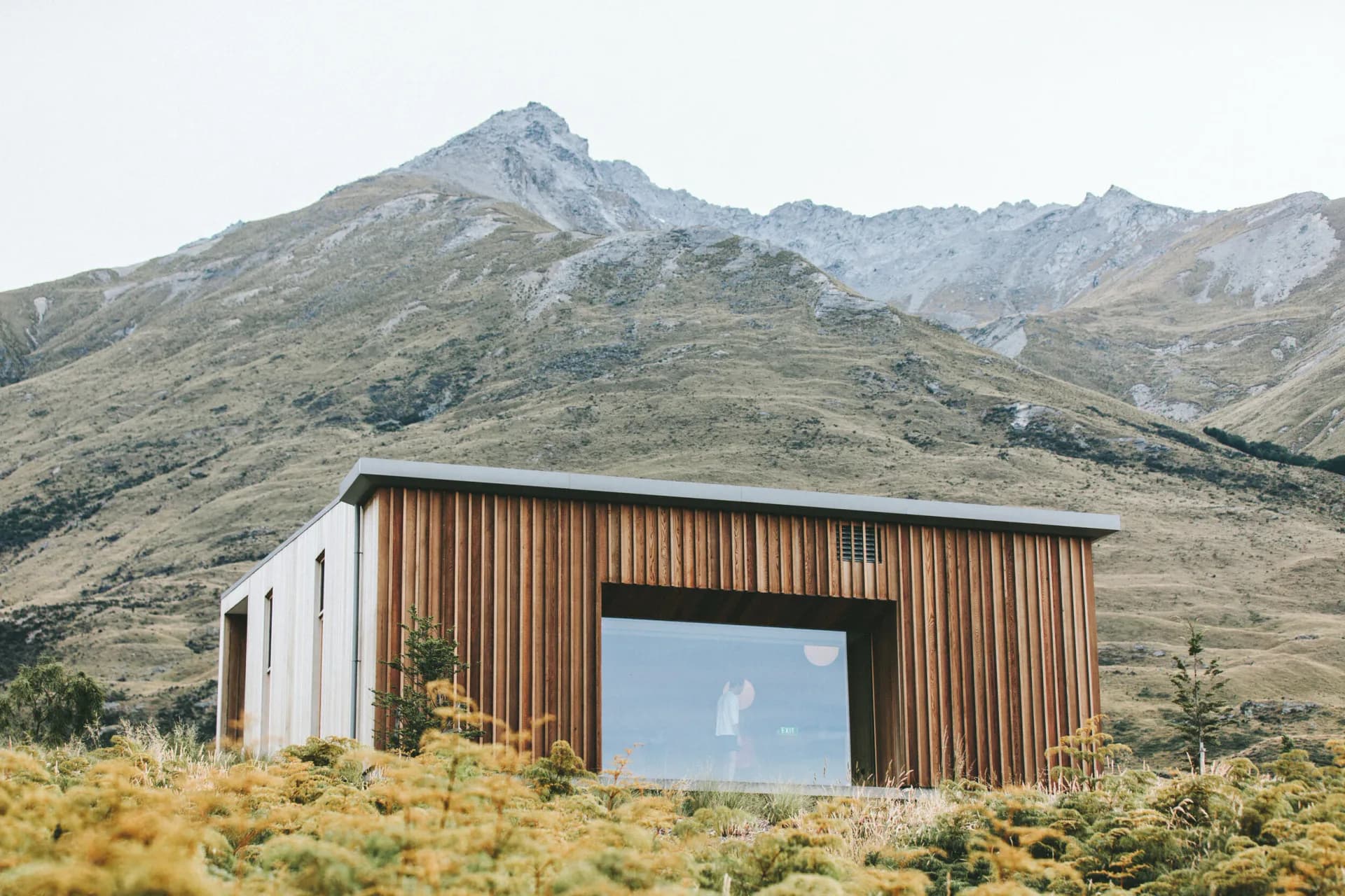 Modern architectural structure with corrugated metal siding and large window opening set in arid mountainous landscape by Illya Azaroff.