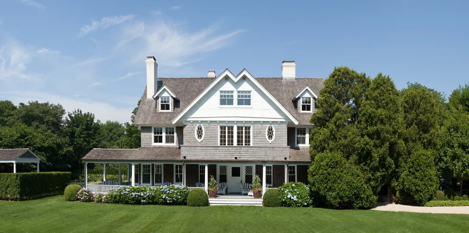 A white colonial-style residential home with symmetrical dormers, black shutters, and a columned front porch overlooking manicured lawns and mature greenery by Inés Elskop.