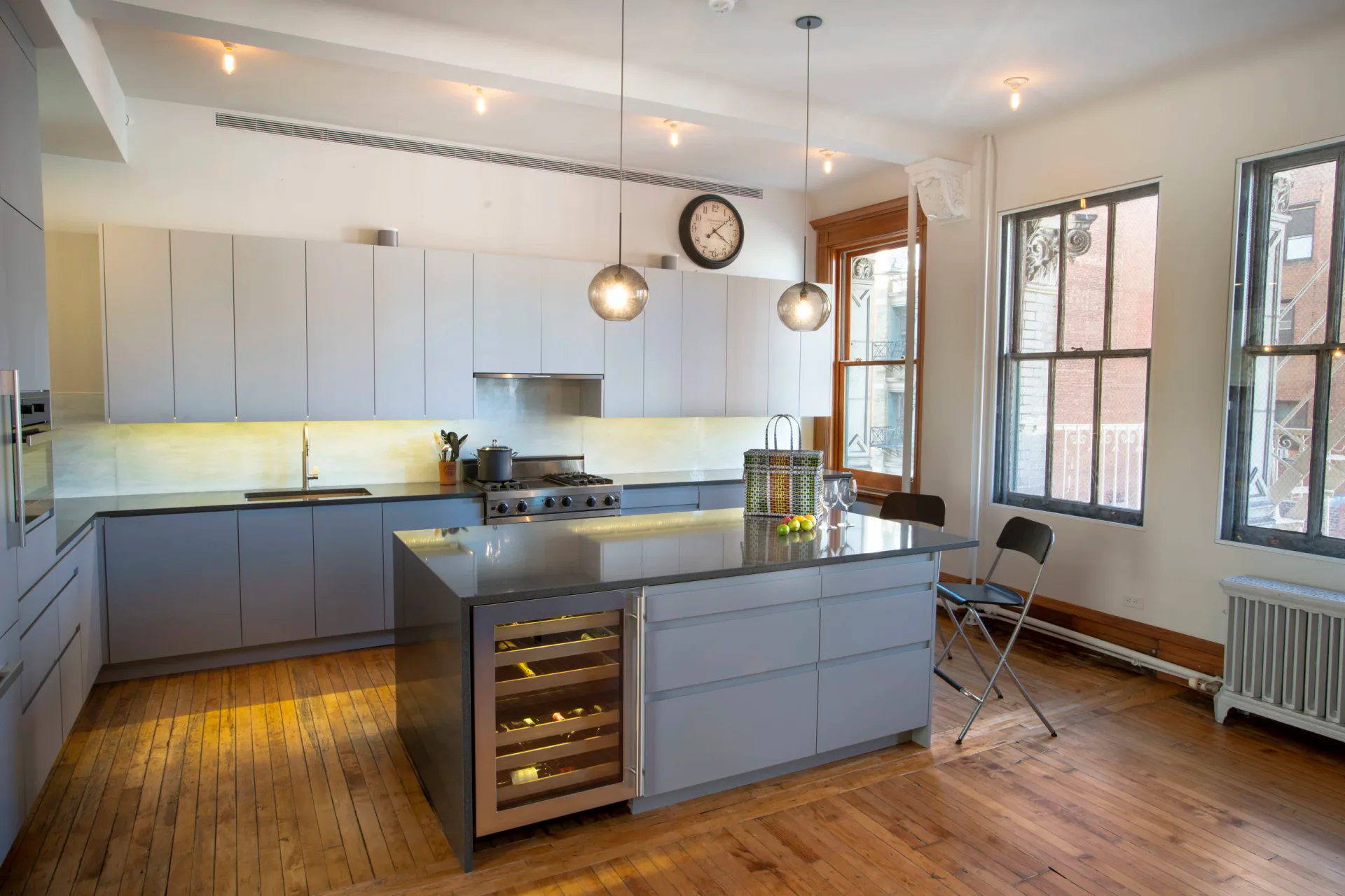 Contemporary kitchen with blue-gray cabinetry, white upper cabinets, stainless steel island with wine cooler, pendant lighting, and warm wood flooring by Inés Elskop.