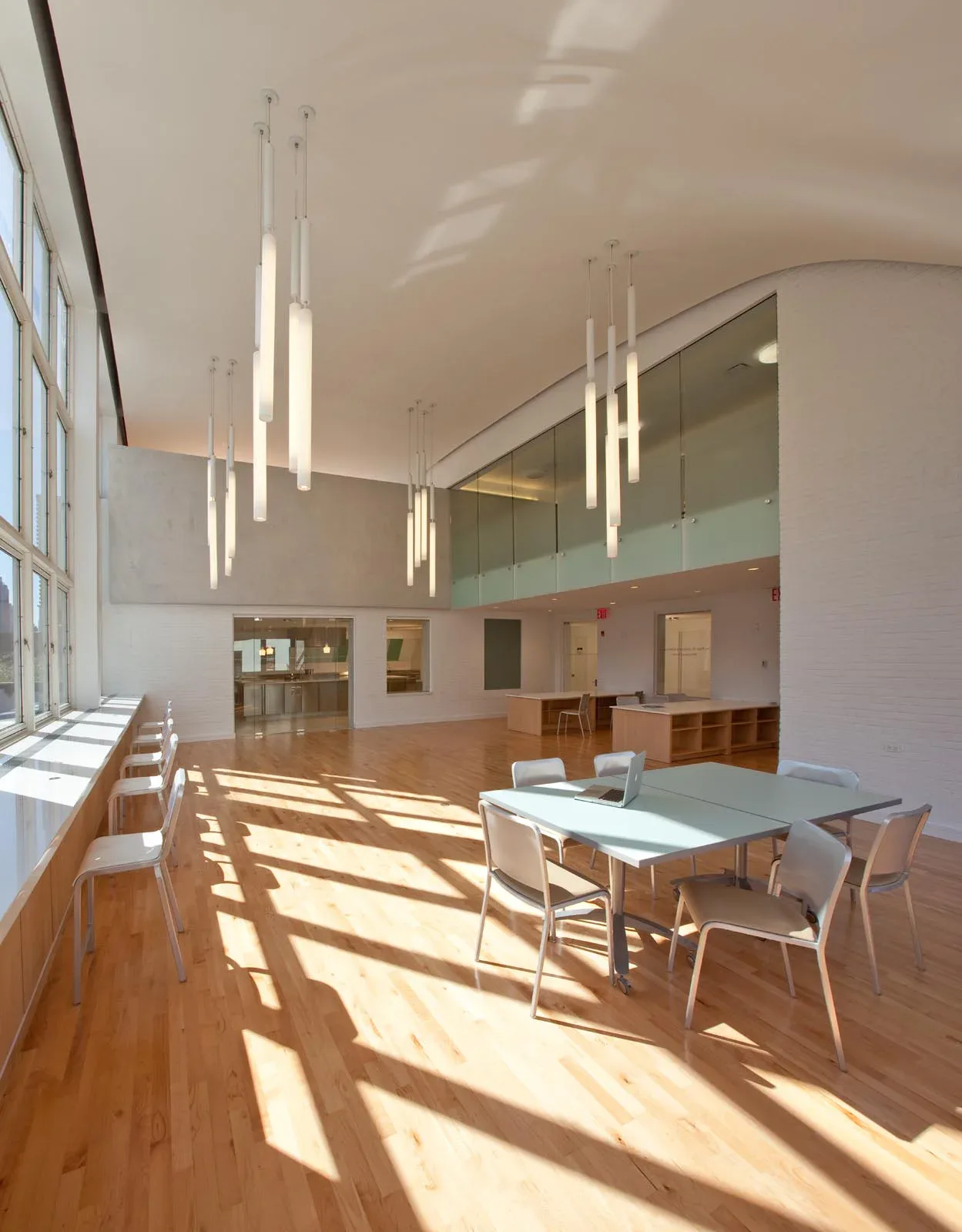 Modern interior with double-height space, wooden flooring, glass mezzanine, pendant lighting, and natural light streaming through large windows by Inés Elskop.