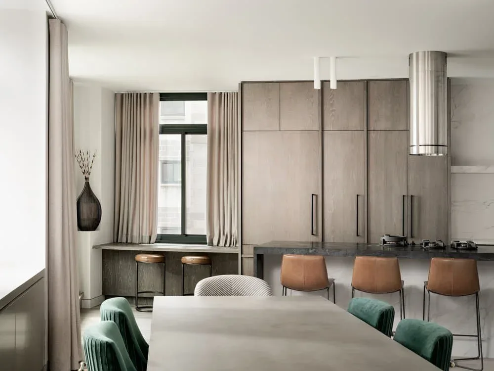 Modern open-plan kitchen and dining space with neutral palette, featuring a white dining table, cognac leather chairs, integrated cooktop island, floor-to-ceiling windows with draped curtains, and minimalist cabinetry by Inga Oren.
