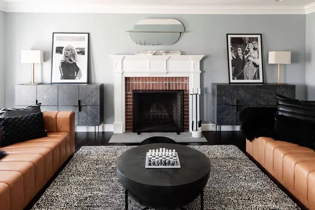 Modern living room with symmetrical cognac leather sofas, black round coffee table, brick fireplace with white surround, gray tile accents, and neutral palette by Inga Oren.