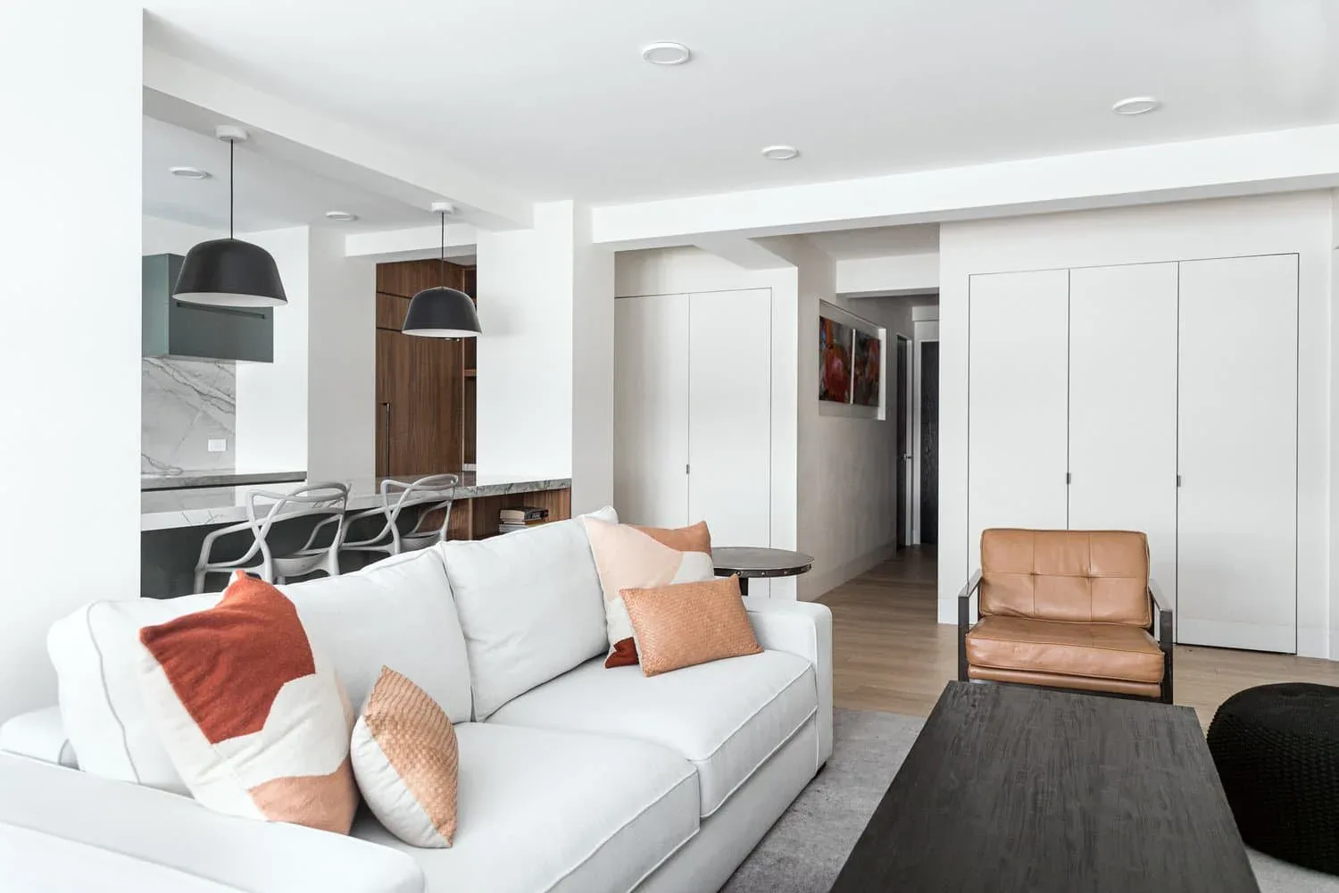 Modern living room with white sofa, warm terracotta accents, open kitchen with dark wood cabinetry, and white built-in closets, designed by Inga Oren.
