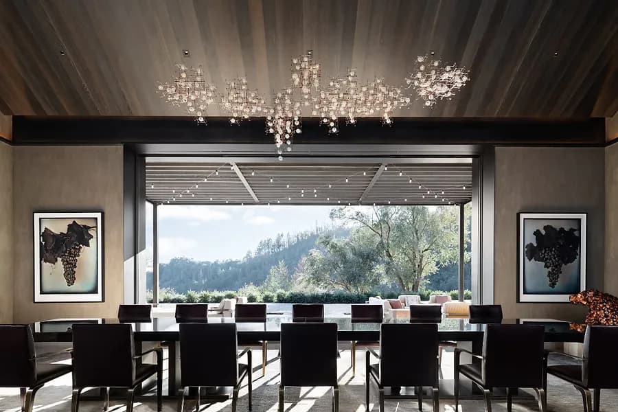 Modern dining room with black chairs, expansive window overlooking forested mountains, crystal chandeliers, and framed artwork by Ingrid Garbar.