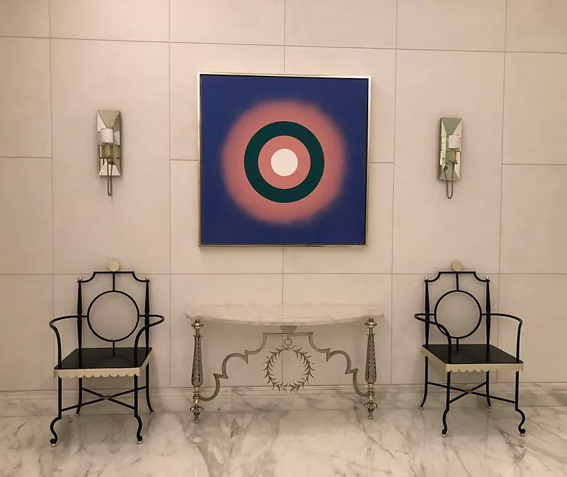 Minimalist interior with cream tile walls, marble floor, symmetrical black metal chairs, central console table, wall sconces, and blue abstract circular artwork by Ingrid Garbar.