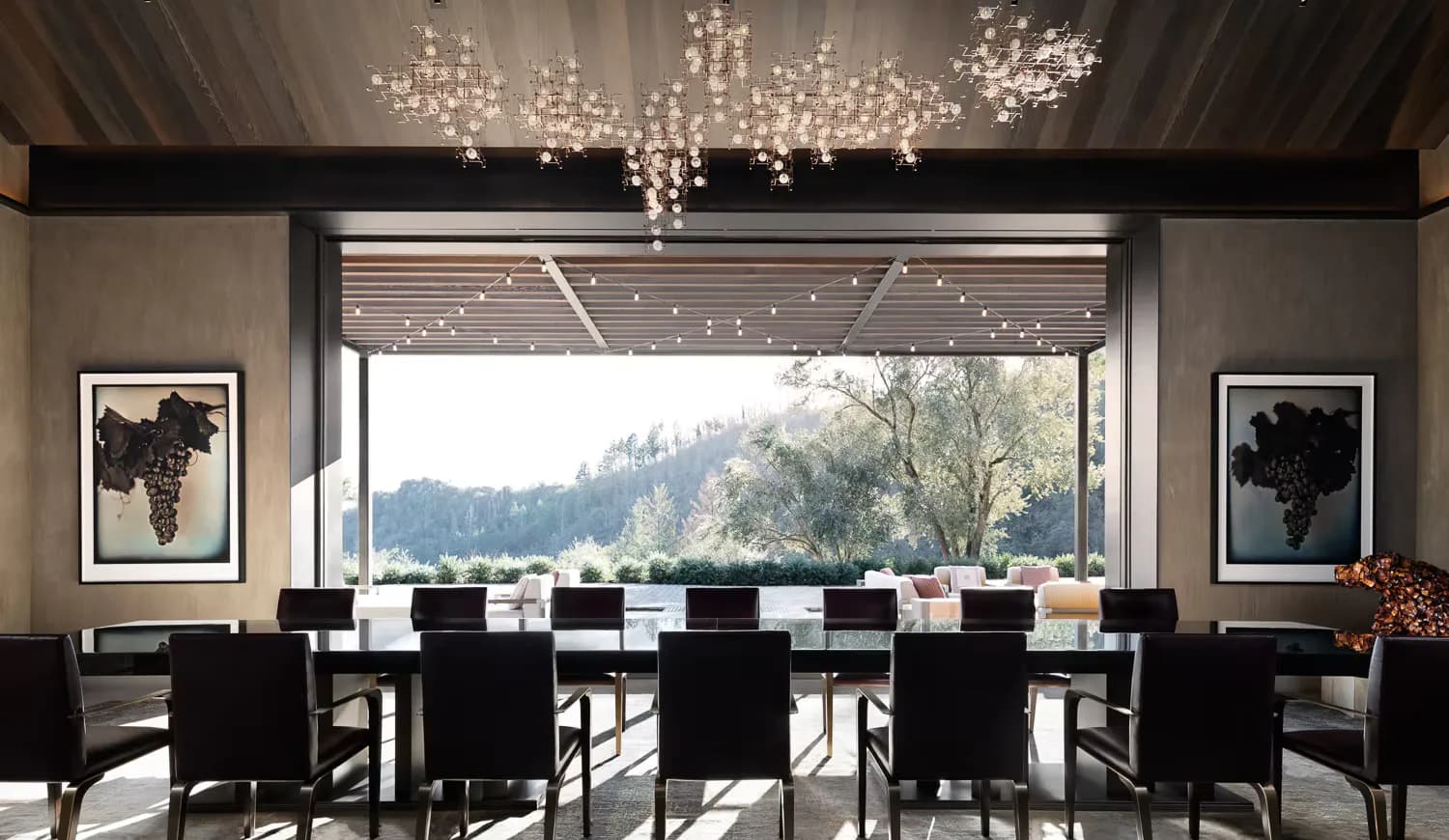 Modern dining room with black chairs, expansive windows overlooking trees, crystal chandeliers, and neutral palette by Ingrid Garbar.