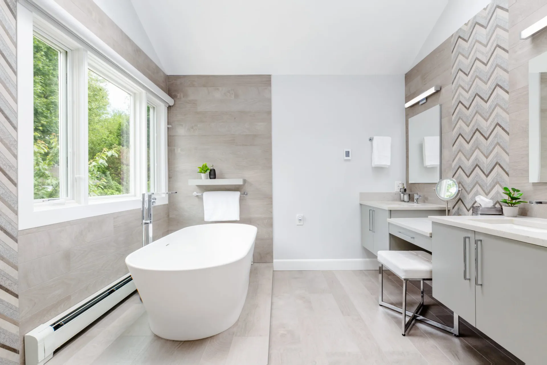 Modern luxury bathroom with freestanding white oval tub, gray tile accents, double vanity with mirror, and natural light from large windows by Ipek Kaypak.