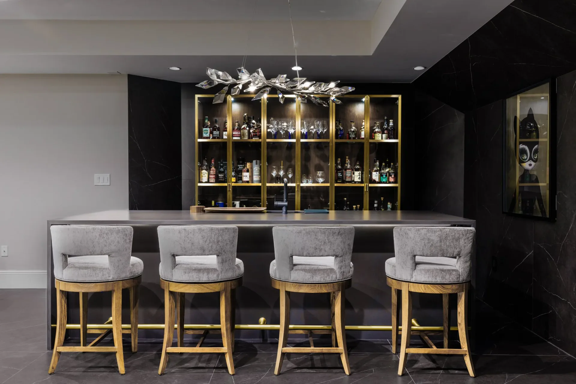 Modern bar interior with four gray upholstered bar stools at a sleek counter, black walls, and illuminated shelving displaying bottles, designed by Ipek Kaypak.