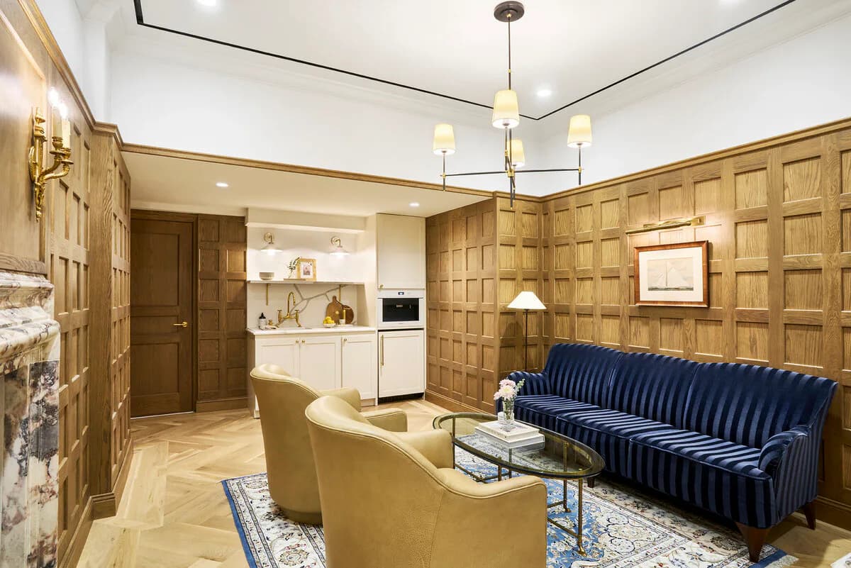 Luxury apartment living room with paneled wood walls, navy striped sofa, brass armchair, and integrated kitchen area by Irma Montefusco.