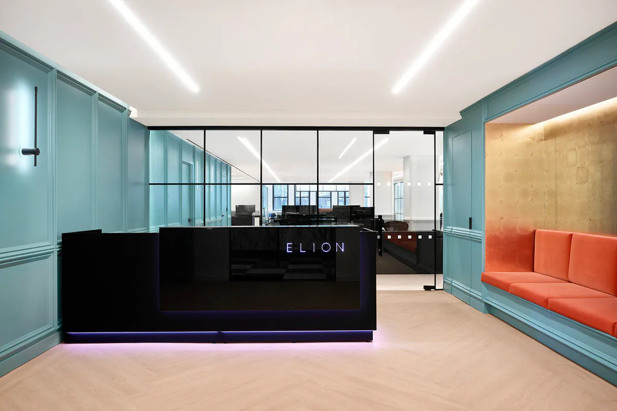 Modern office reception area with turquoise walls, black reception desk with illuminated 'ELION' signage, coral seating, and glass partitions by Irma Montefusco.