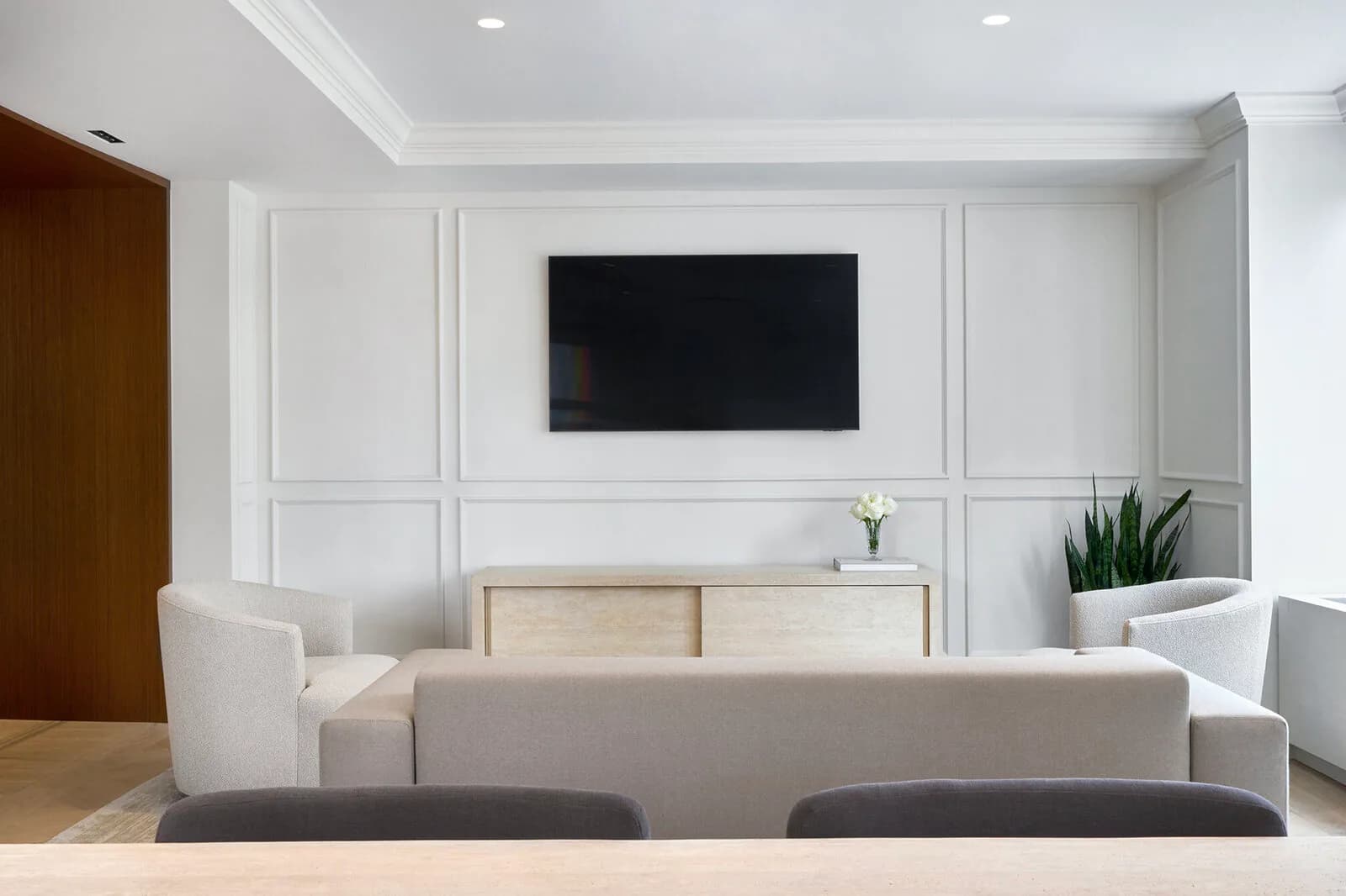 Modern living room with cream sofa, light wood media console, white paneled walls, mounted TV, and potted plants in a minimalist interior by Irma Montefusco.