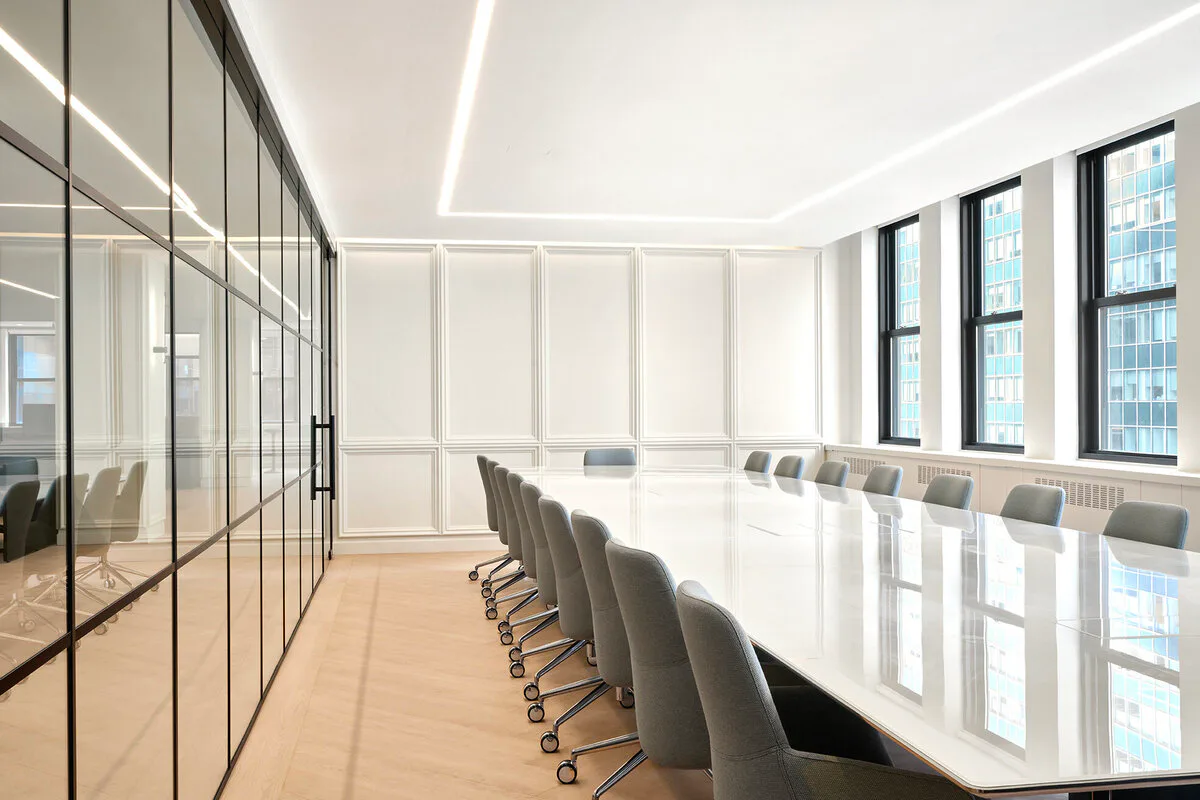 Modern conference room with floor-to-ceiling glass partitions, long white table, gray chairs, and floor-to-ceiling windows overlooking a city skyline, designed by Irma Montefusco.