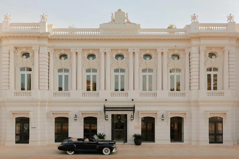 Neoclassical cream-colored facade with symmetrical arched windows, ornamental balustrade, and dark vintage car parked in front by Isaac Jimenez.