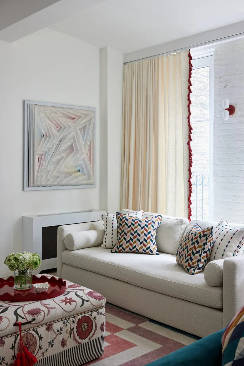 Modern living room with cream sofa, patterned pillows, fireplace, large windows with cream curtains, and red decorative accents by Isabel Runsewe.