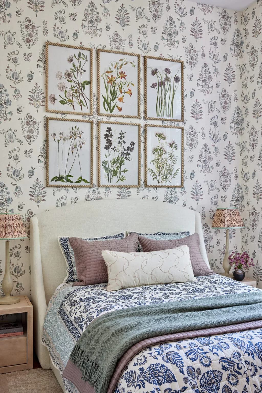 Bedroom interior with botanical framed prints above upholstered headboard, floral wallpaper, layered bedding in blue and mauve tones, and natural wood nightstands by Isabel Runsewe.