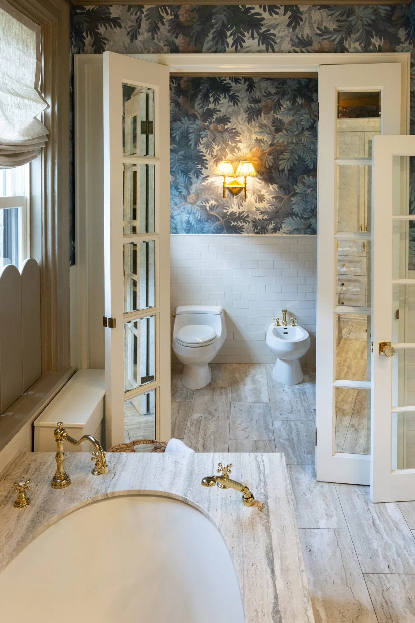 Luxurious master bathroom with marble flooring, brass fixtures, wallpapered accent wall with fern motif, and adjoining toilet room with sconce lighting, designed by Isabel Runsewe.