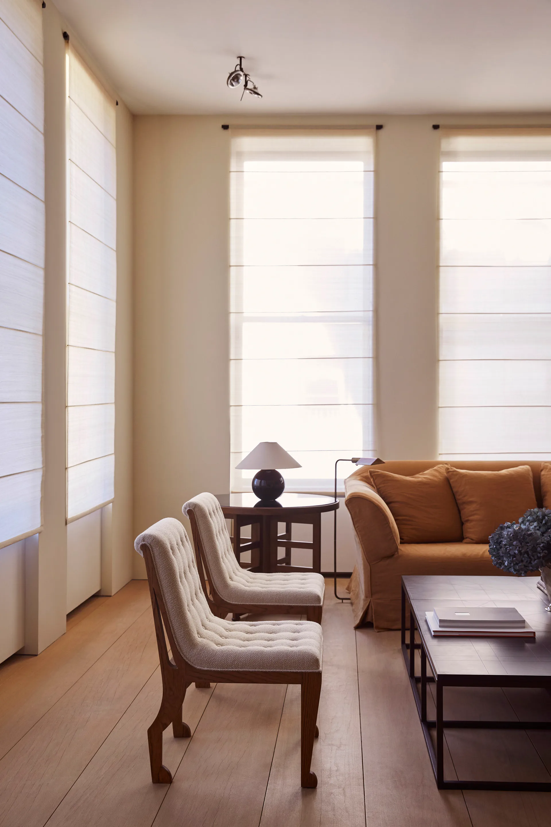 Bright, minimalist living room with tall windows, cream and tan palette, upholstered chairs, caramel sofa, and dark wood accents by Isabella Nuelle.
