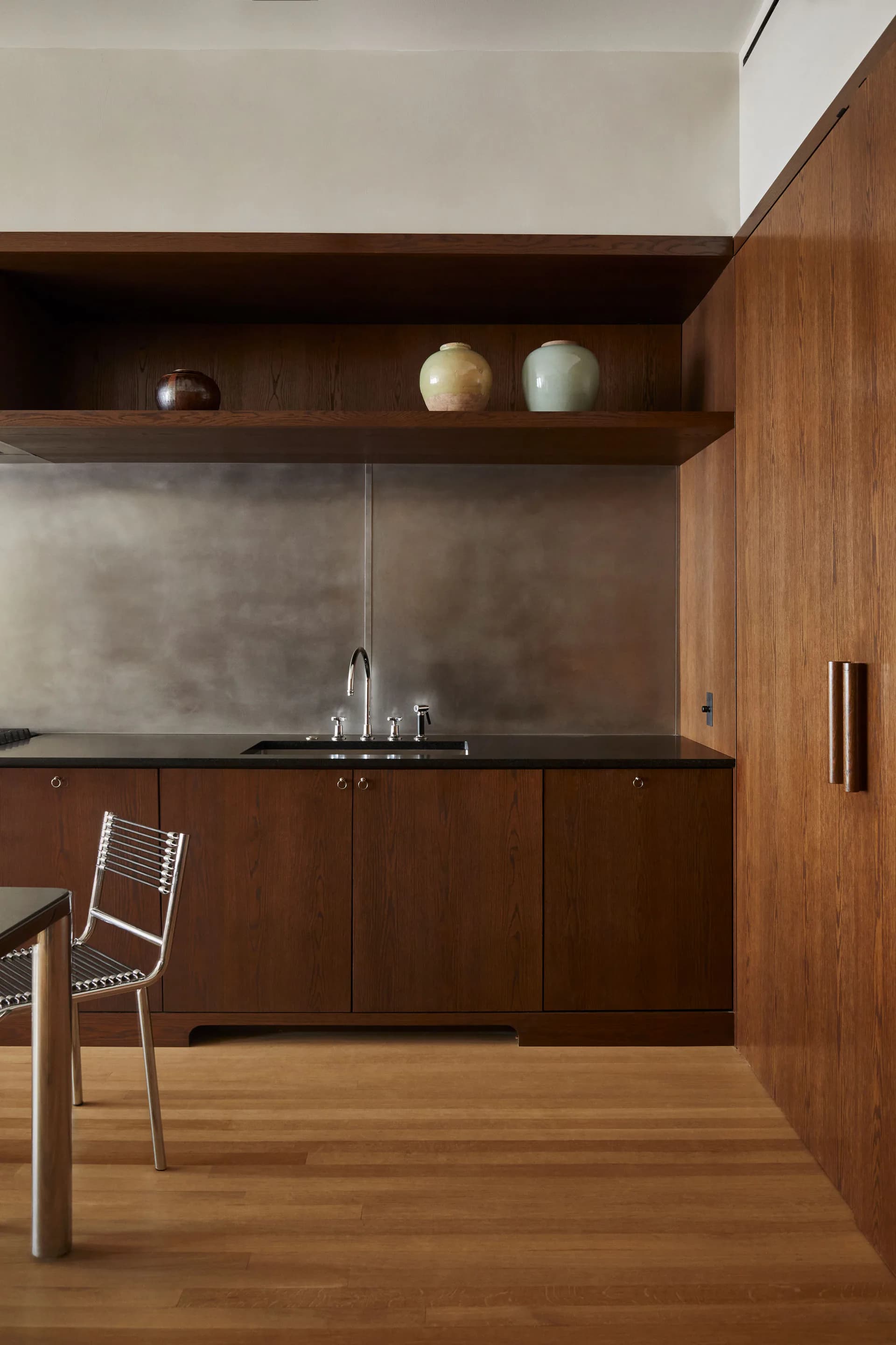 Modern kitchen with warm wood cabinetry, concrete backsplash, floating shelf with decorative vessels, and wooden flooring by Isabella Nuelle.