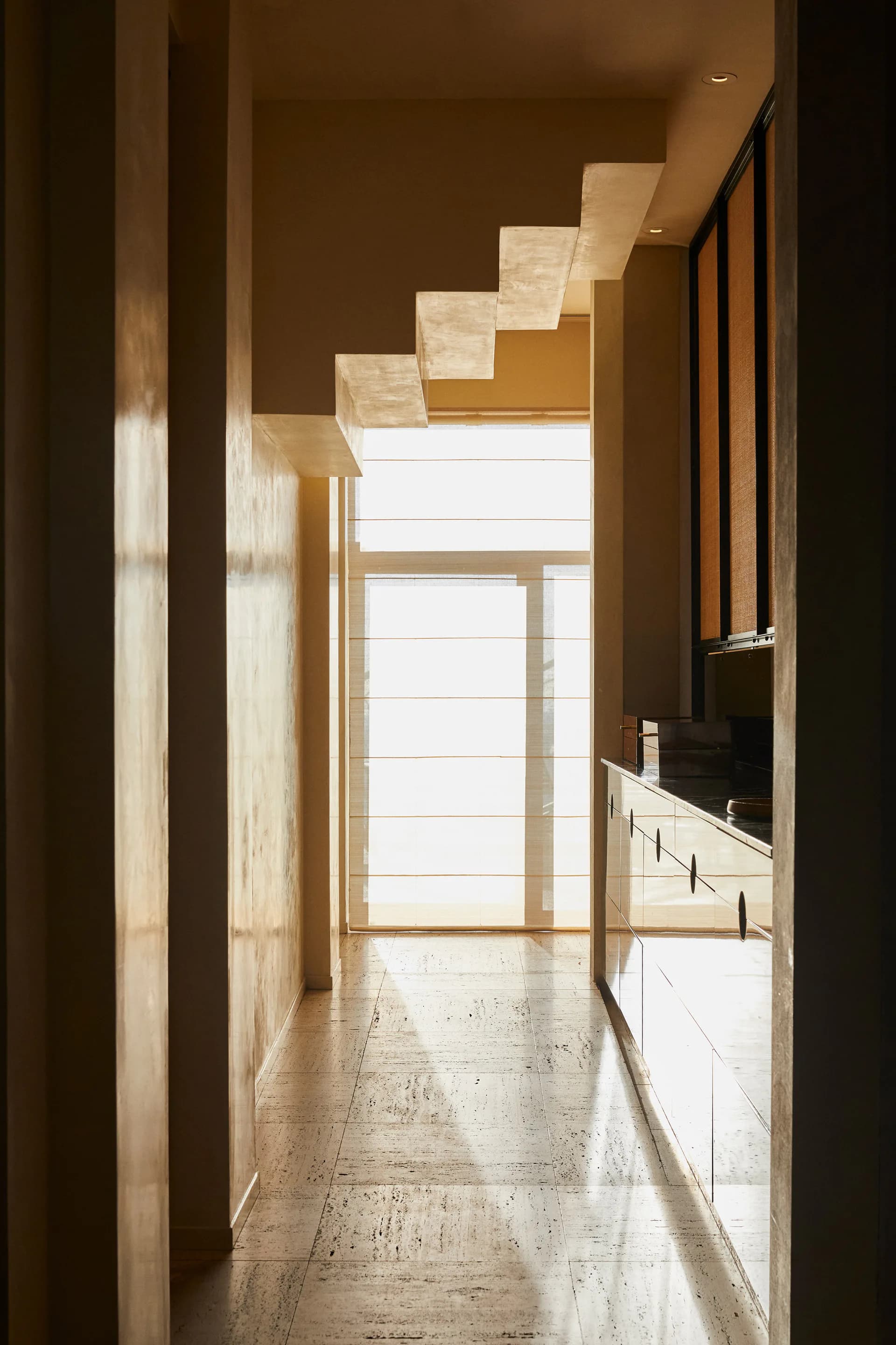 Modern hallway with warm beige stone flooring, brown walls, and geometric skylight arrangement creating angular light patterns, by Isabella Nuelle.