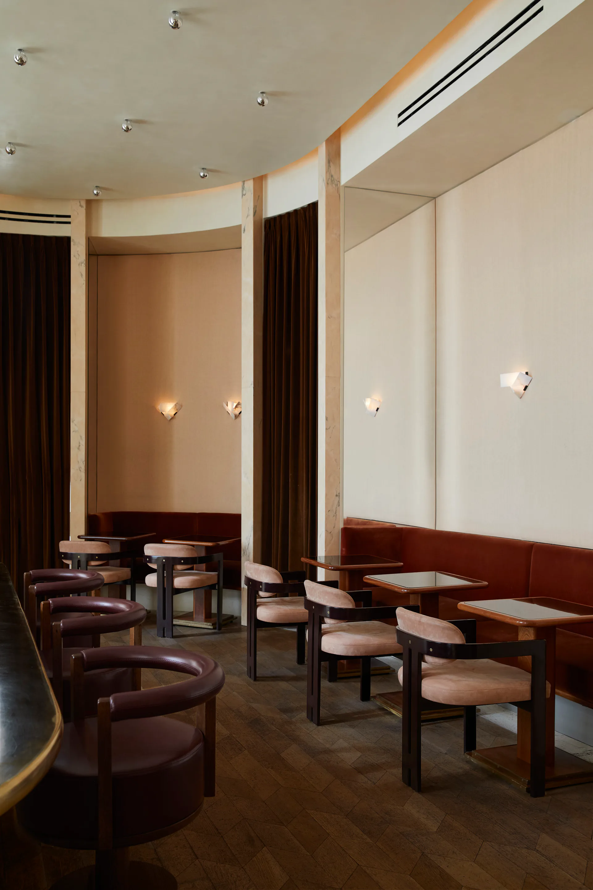 Contemporary lounge interior with curved burgundy leather seating, wood-framed alcoves, warm beige walls, and sculptural wall sconces by Isabella Nuelle.
