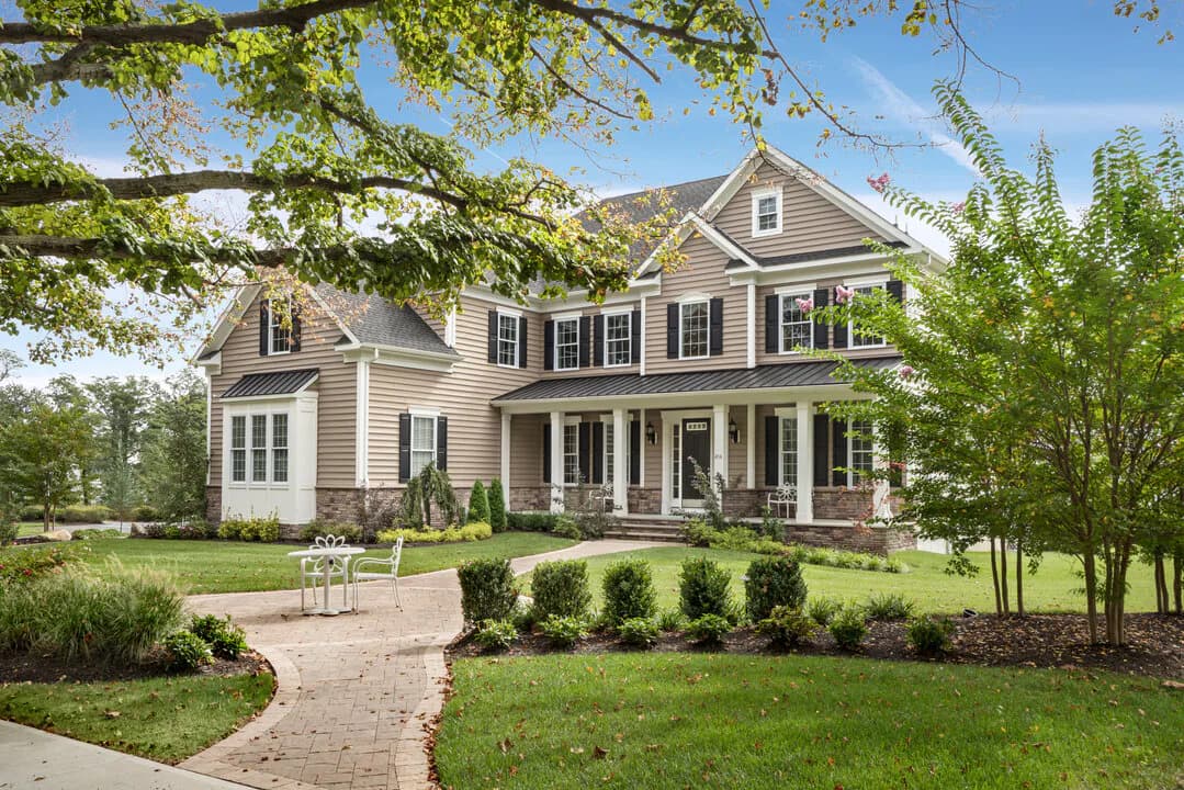 A two-story farmhouse with gray-taupe siding, white trim, and black shutters sits on manicured grounds with brick walkway and landscaping by Jack Murphy.