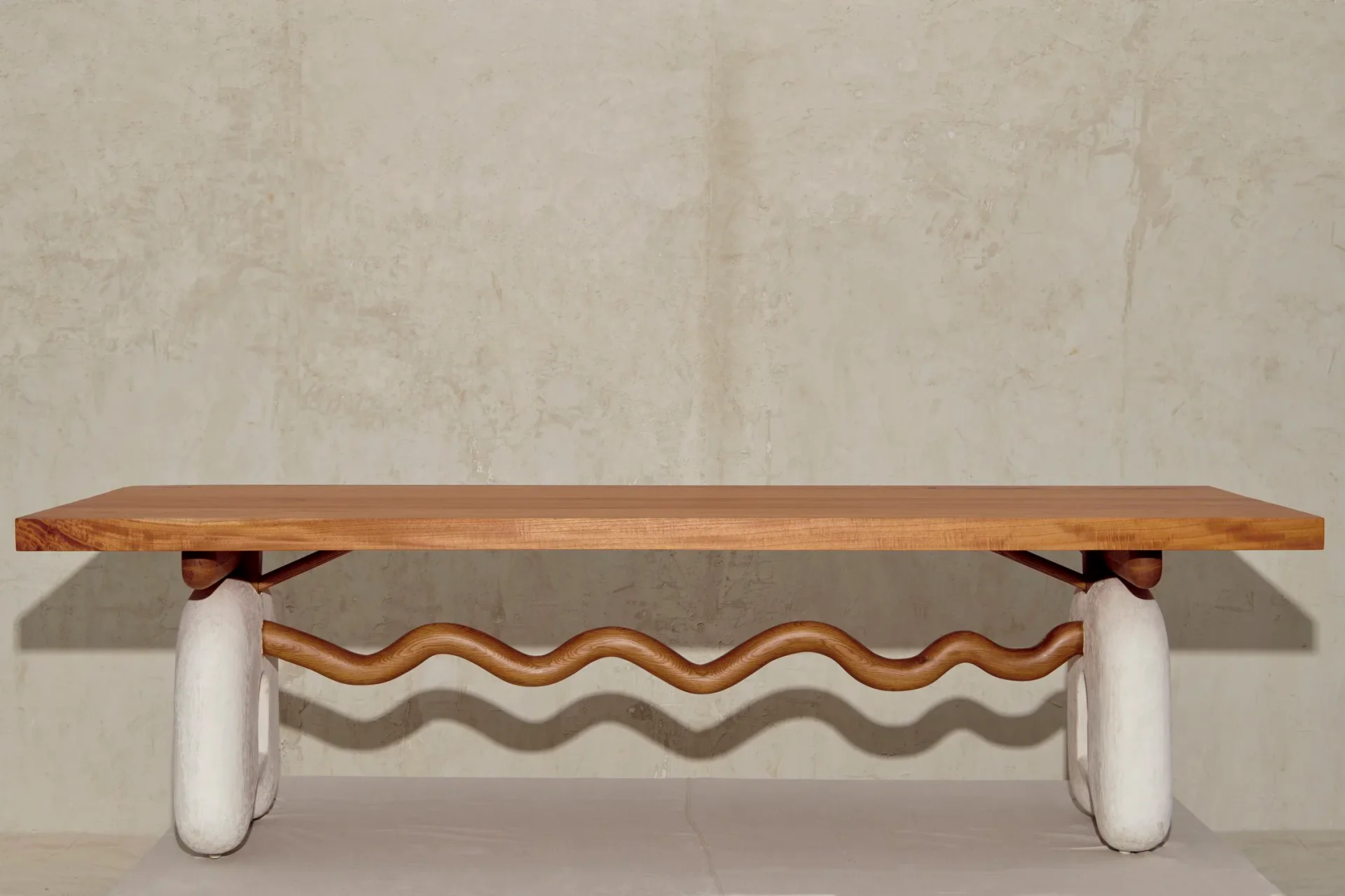 Sculptural wooden bench with wavy decorative support in warm wood tones and white cylindrical legs against neutral wall by Jackrabbit Studio.