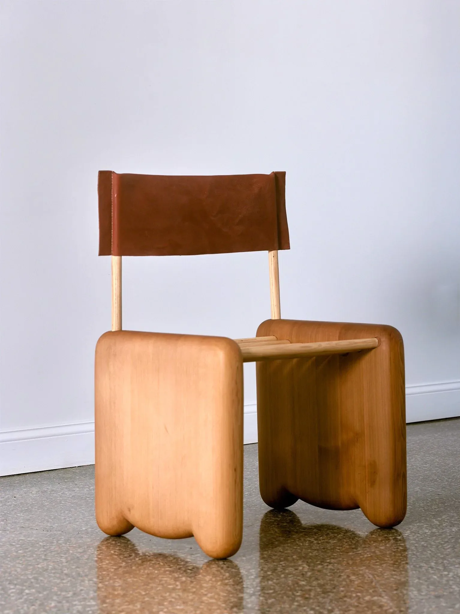 Sculptural wooden chair with curved organic forms in warm honey tones, featuring a brown leather backrest, photographed in a gallery setting by Jackrabbit Studio.