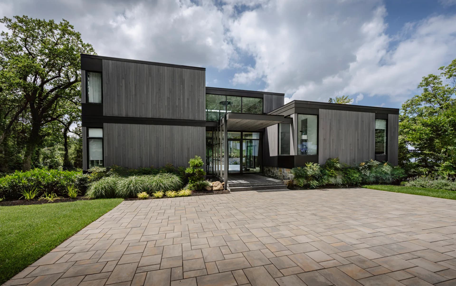 Modern black-clad residential home with glass elements and natural landscaping, featuring a wide paver driveway by Jaclyn Letsch.