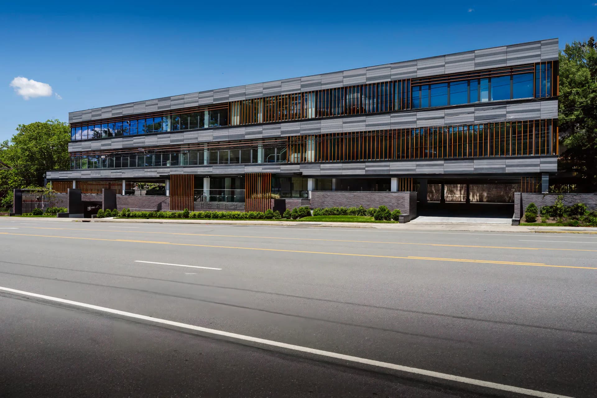 Modern three-story commercial building with horizontal wood and metal slatted facade, expansive glazing, and clean landscaping along street frontage by Jaclyn Letsch.