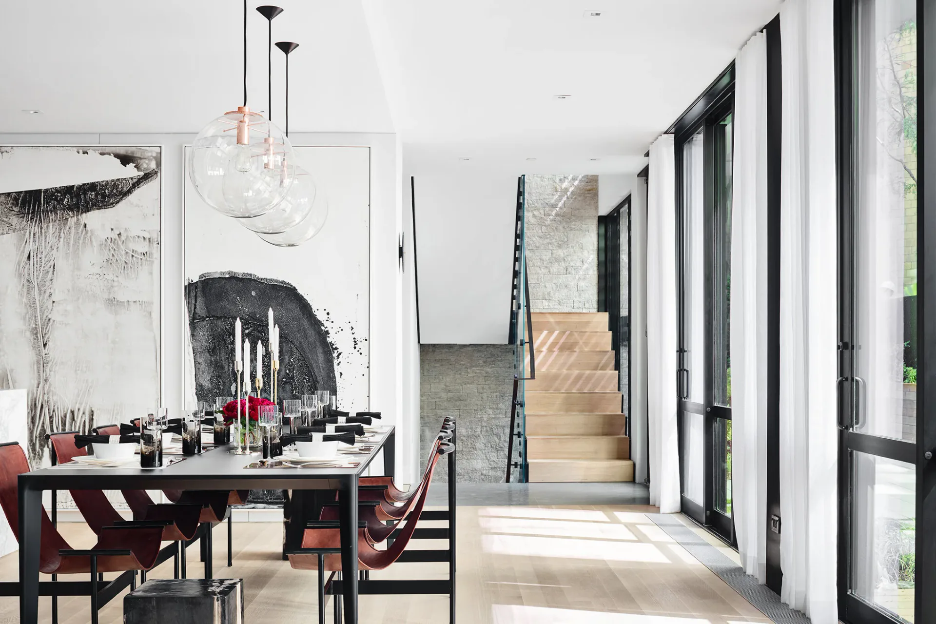 Modern dining space with black dining table, burgundy chairs, and abstract artwork, connected to a bright corridor with large windows and natural wood flooring, by Jacqueline Nguyen.