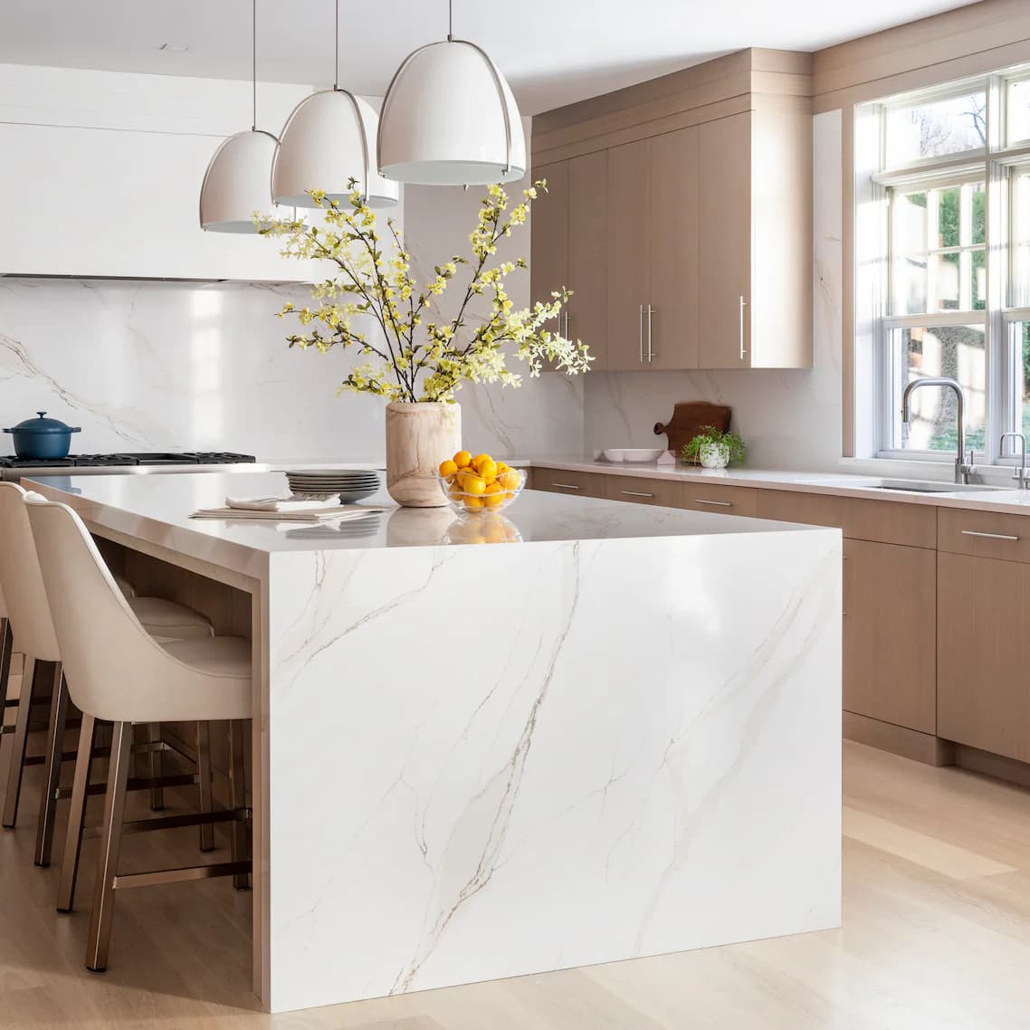 Modern kitchen with white marble island, natural wood cabinetry, pendant lighting, and yellow flowers by Jacquelyn Lagravinese.