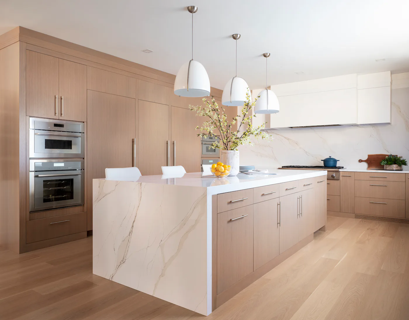 Modern kitchen with light wood cabinetry, white marble island, stainless steel appliances, and pendant lighting by Jacquelyn Lagravinese.