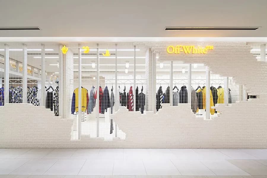 Modern retail storefront with white framework displaying colorful jackets and apparel, yellow neon signage, and minimalist interior design by James Wines.