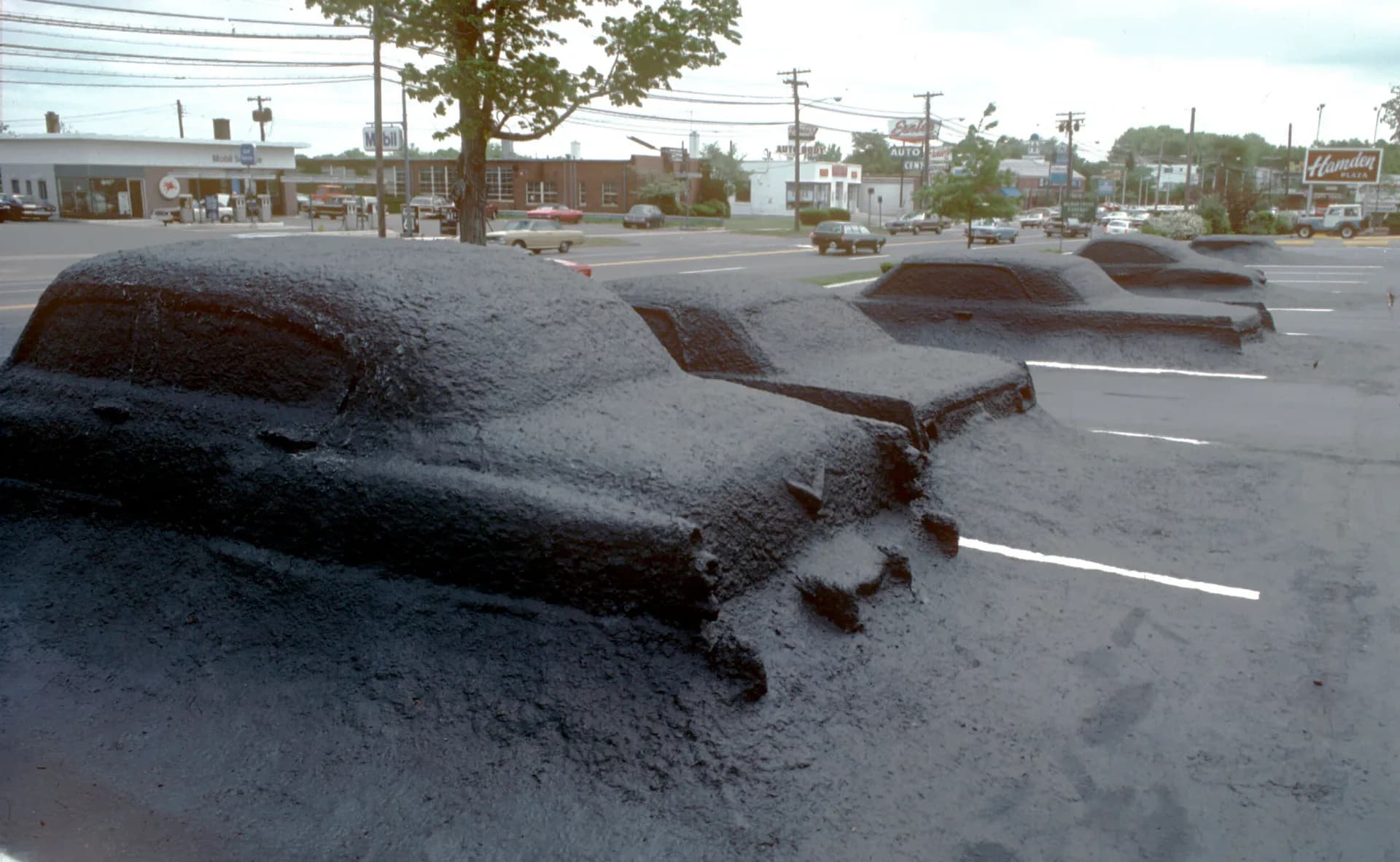 Sculptural concrete forms resembling whale shapes installed in a parking lot, with urban streetscape and buildings visible in the background, by James Wines.