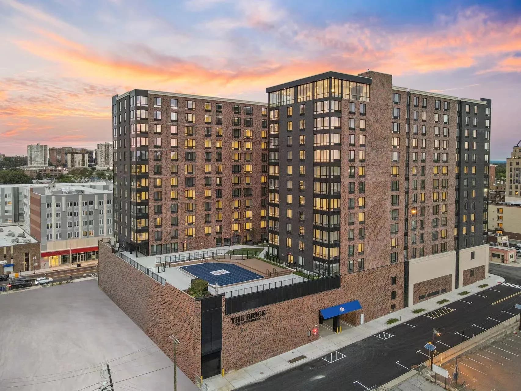 Modern brick residential tower with illuminated windows at dusk, featuring a courtyard pool and surrounding urban landscape by Jasmine Jia.