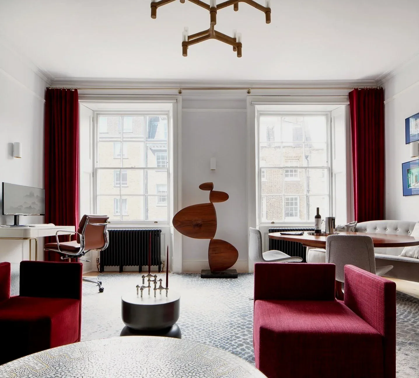 Modern living room with burgundy upholstered furniture, wooden sculptural art piece, large windows with red curtains, and minimalist accessories by Jasmine Lam.